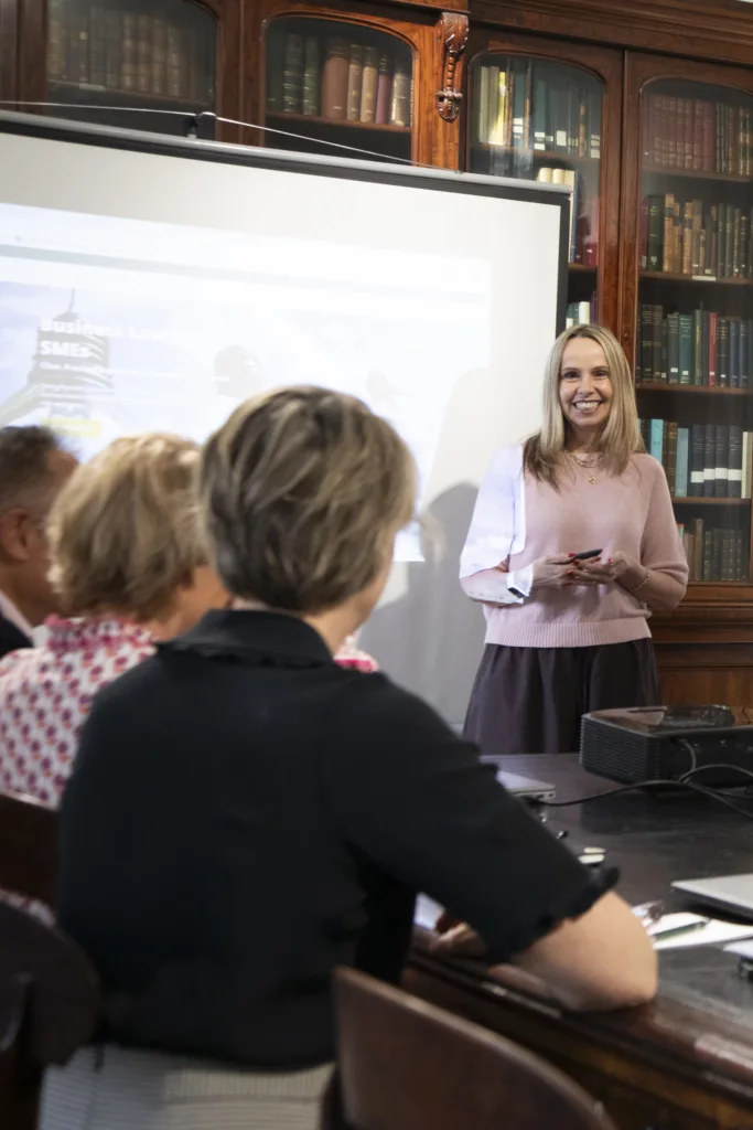 A woman stood beside the projector screen, leading the meeting while participants seated around the meeting room table listened attentively and actively engaged in the discussion.
